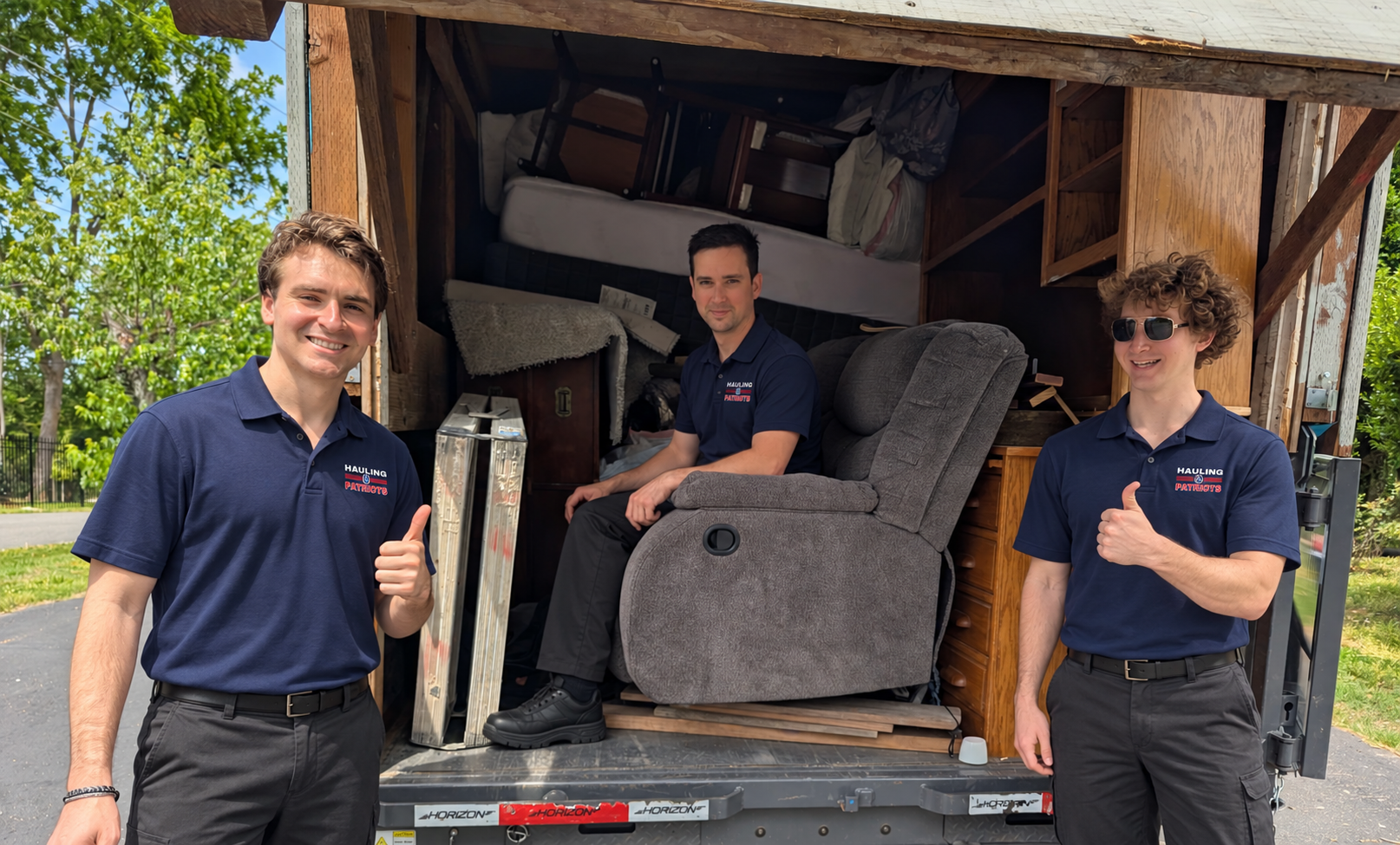 The Hauling Patriots crew in uniform giving a thumbs up at a loaded moving trailer with a recliner and furniture